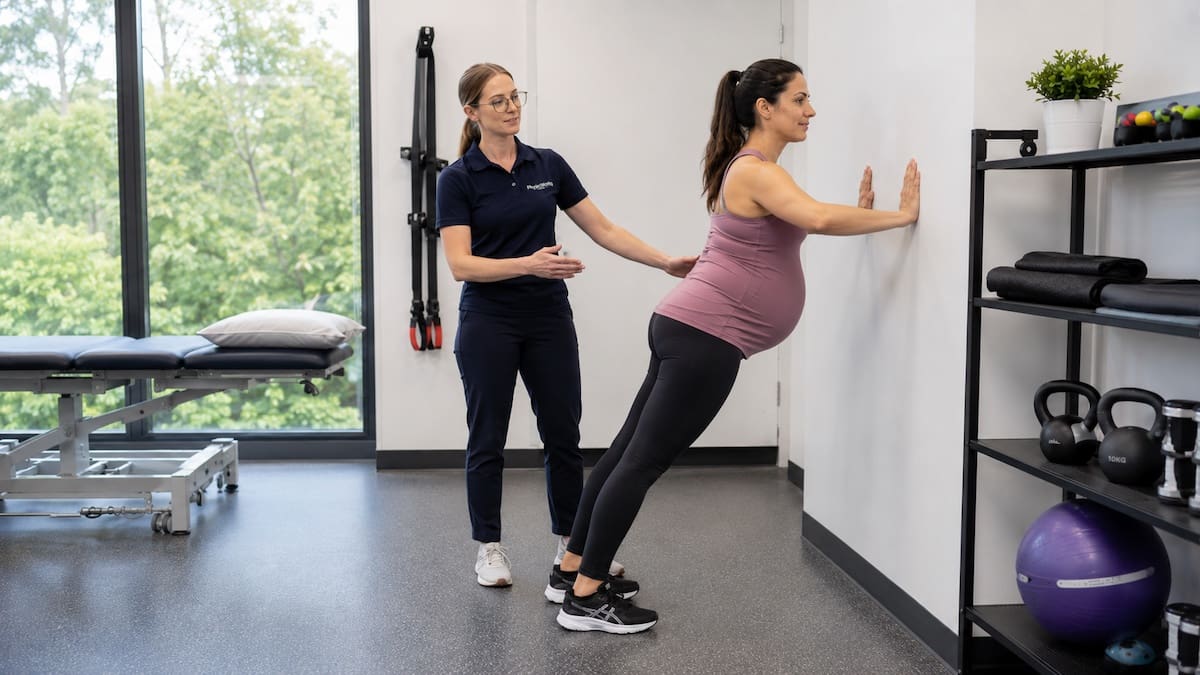 Pregnant woman performing a wall push-up while a physiotherapist observes her technique
